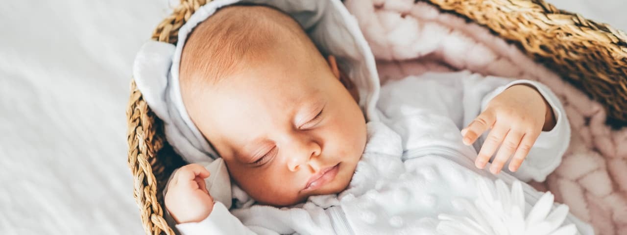 A baby dressed in white peacefully sleeping in a wicker crib.