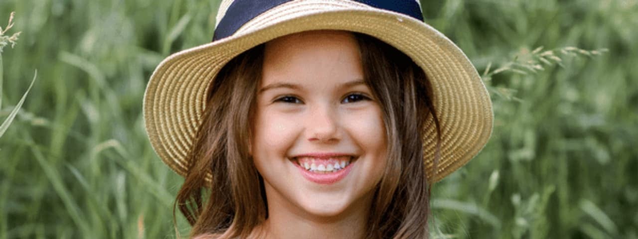 A girl in a straw hat smiling in a grassy field.
