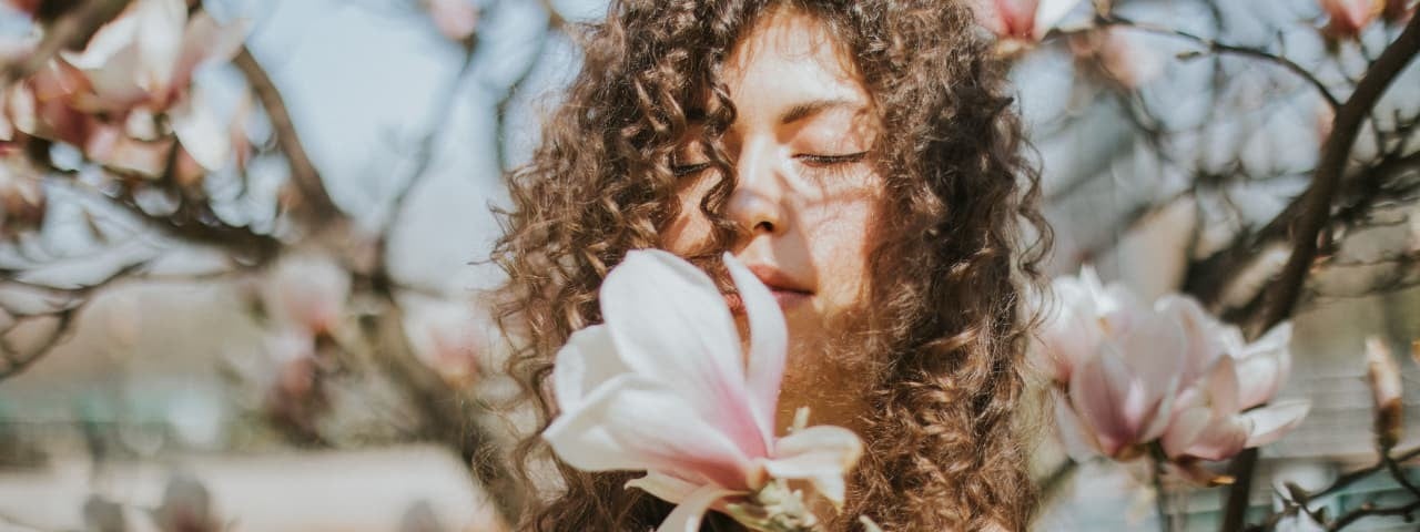 A curly-haired girl leaning in to smell a flower on a tree branch.