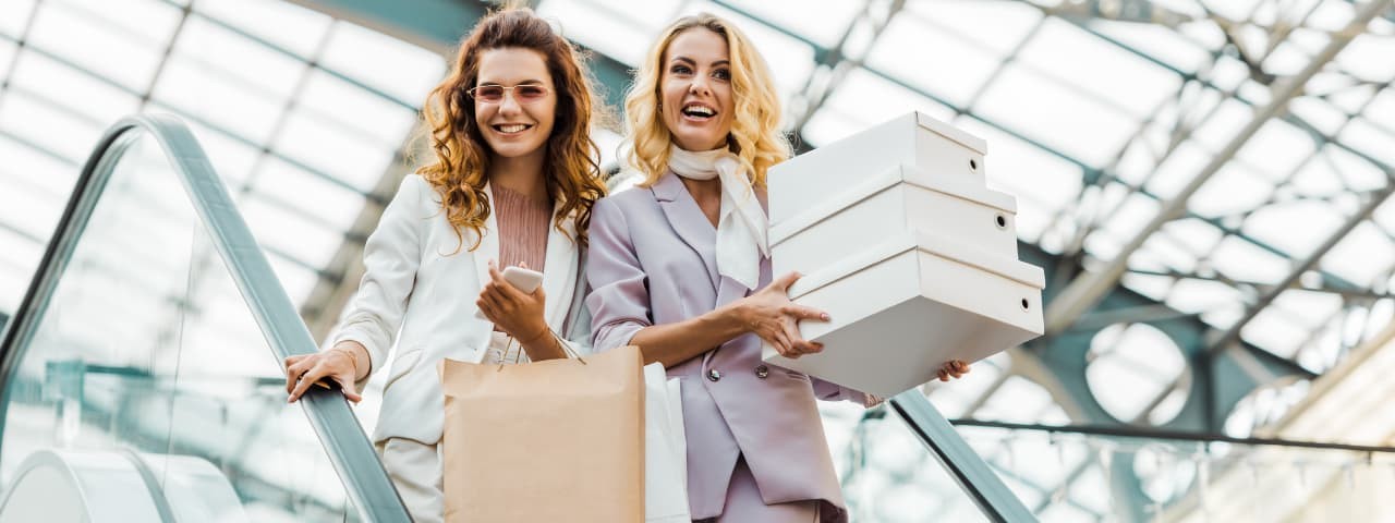 Two girls at the top of an escalator, carrying shopping bags and boxes.
