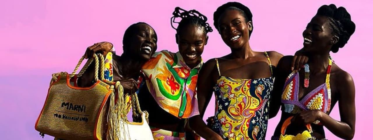 Four women in vibrant dresses and handbags posing in front of a beach backdrop.